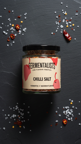 Jar of Fermentalists Chilli Salt on a dark surface with scattered chili peppers and salt crystals.